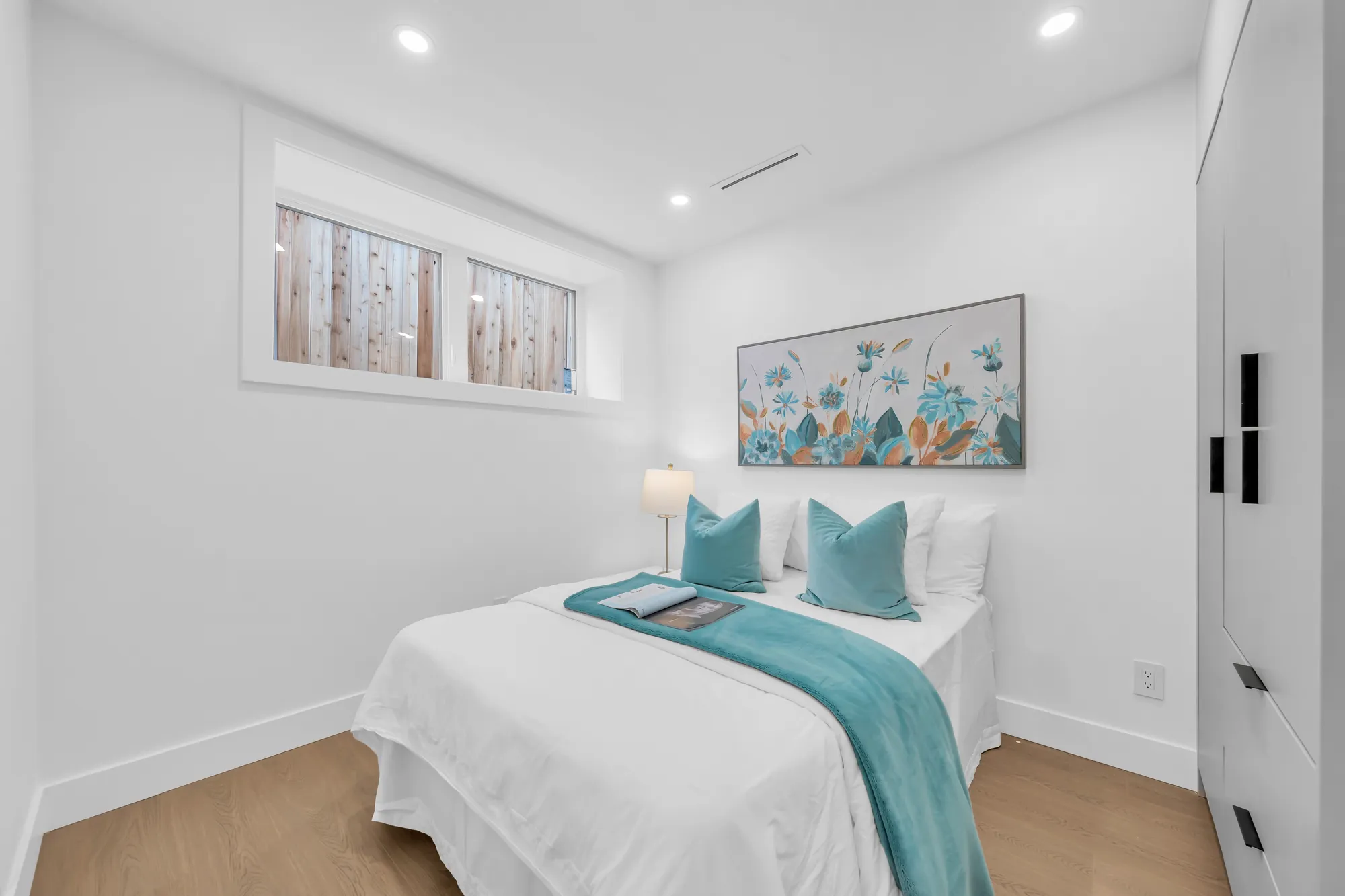 A bedroom: a bed with white bedding and a teal throw, a framed print, windows set high in the wall