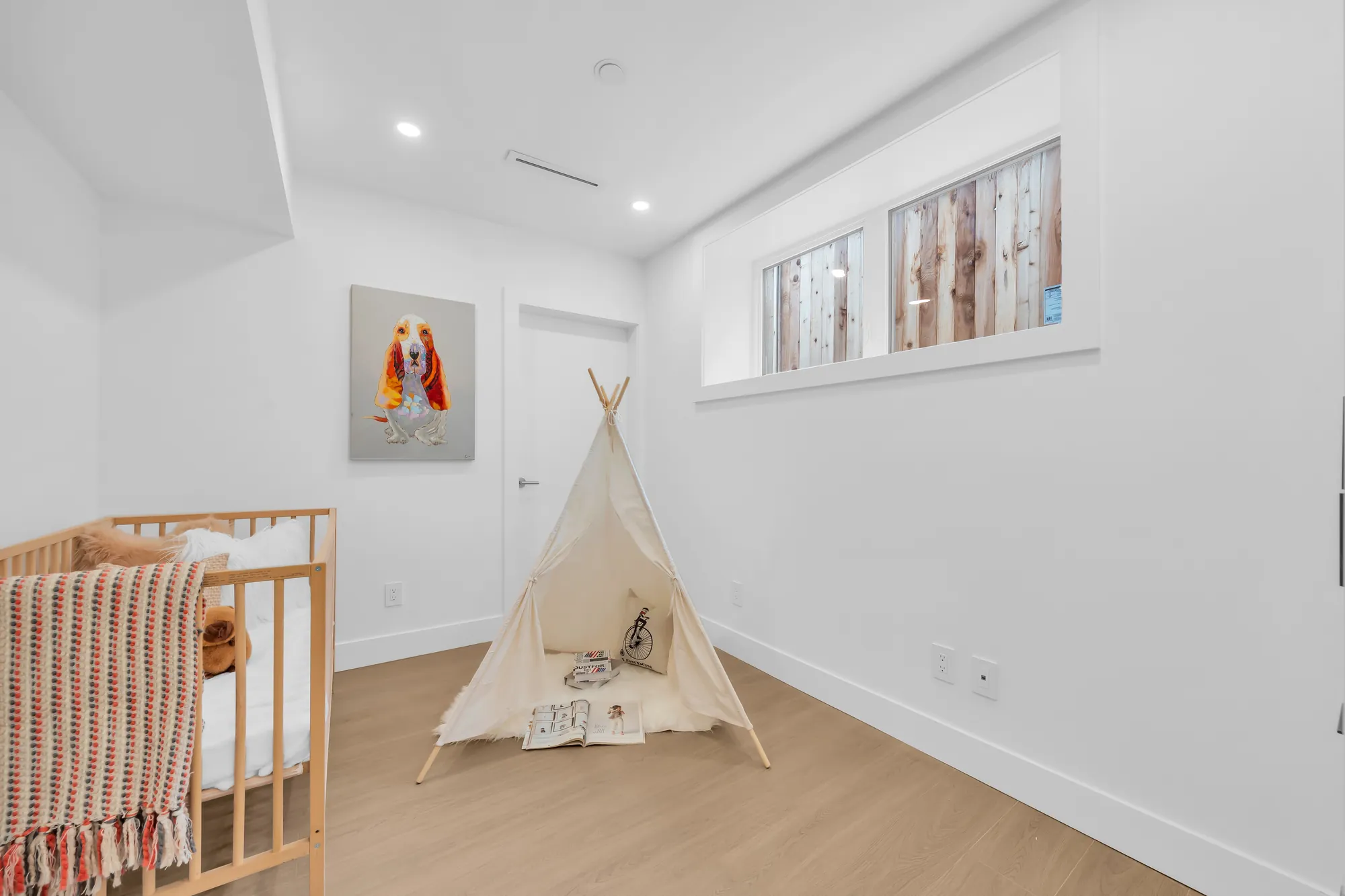A room set up as a nursery: a wood crib, a play tent, a framed print, a window set high
