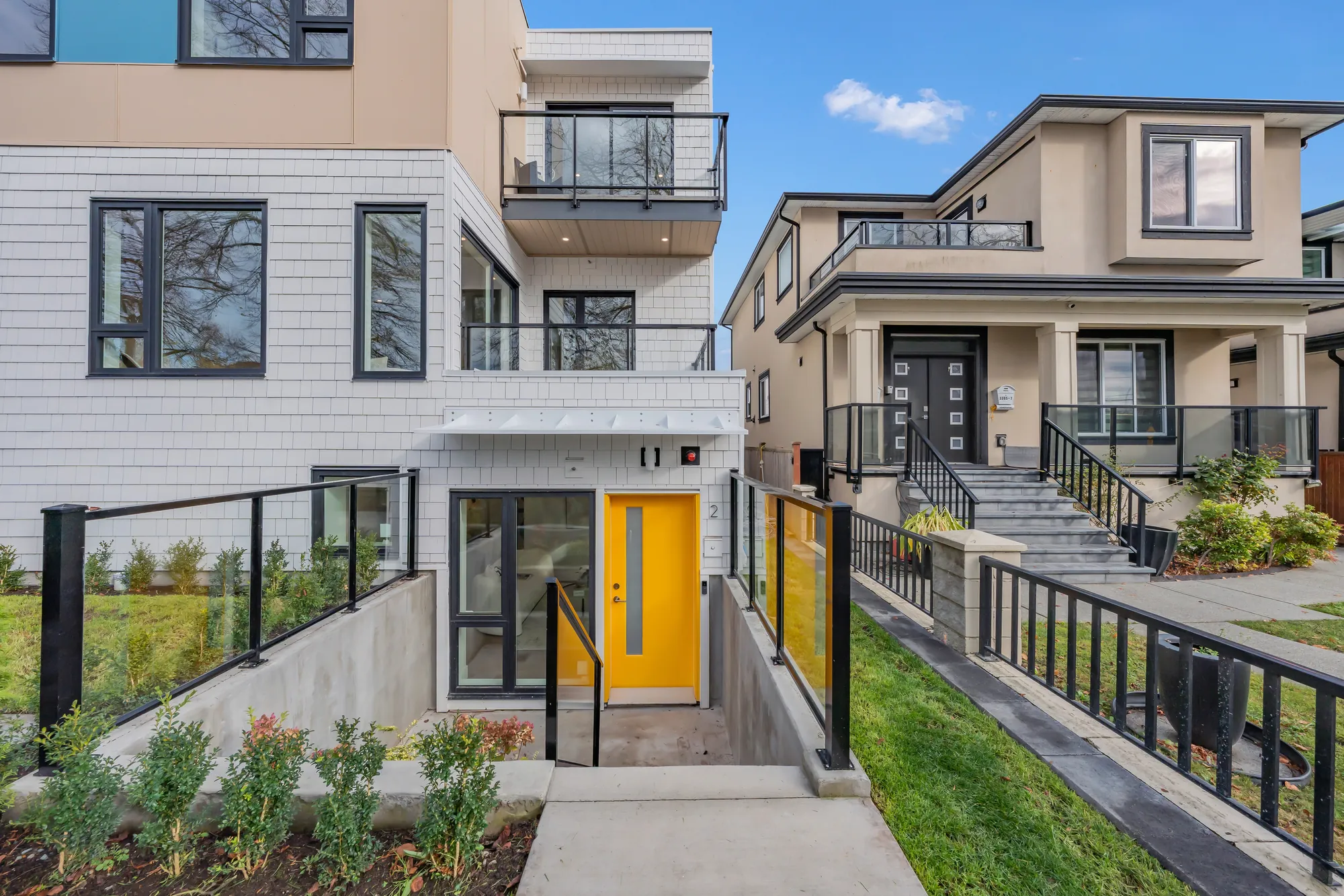 The front door of 2-3391 Price Street: a yellow door and a wide window at the bottom of a sunken patio with glass and steel railings, under the white shingle siding of the new building