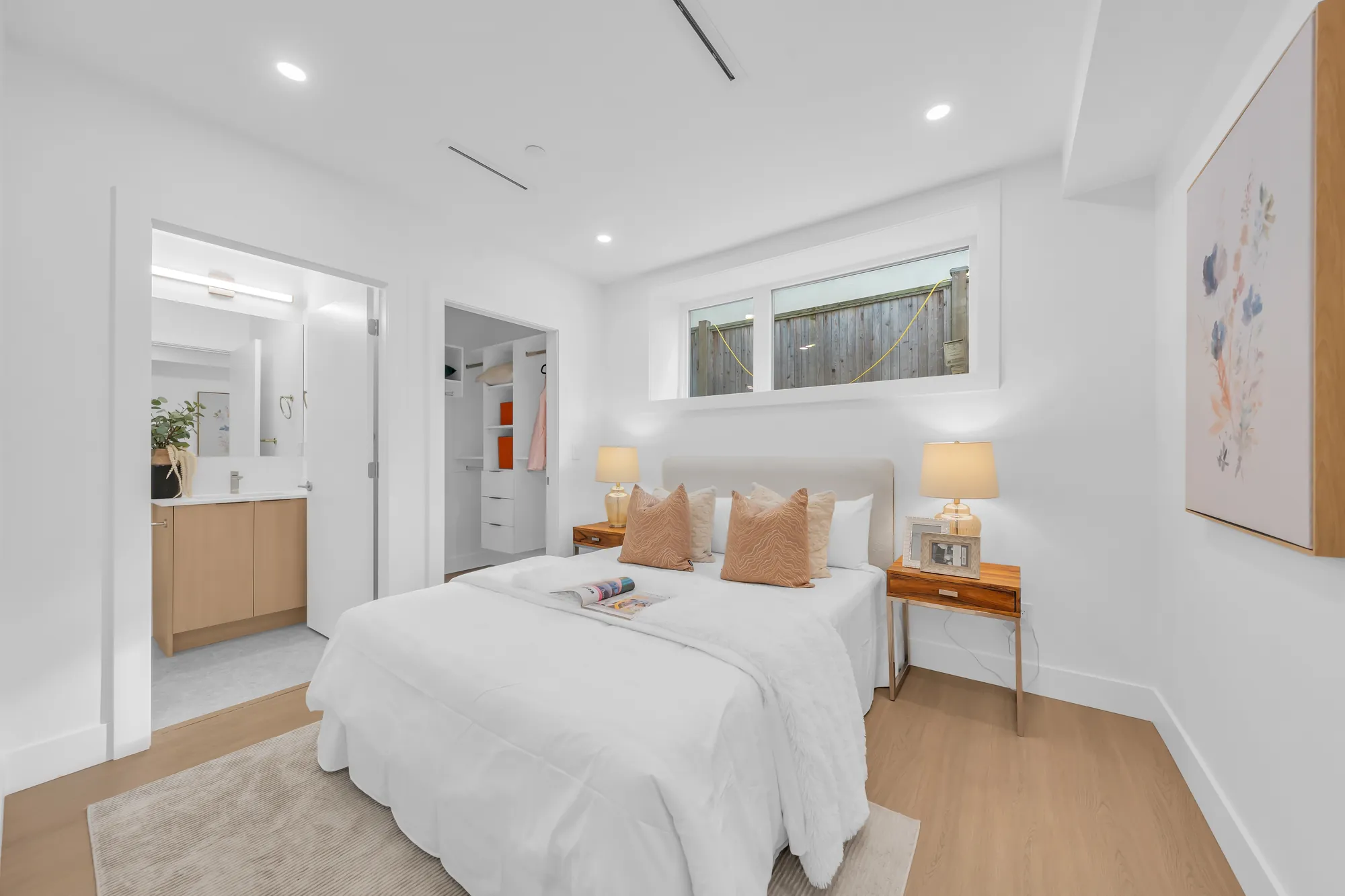 The primary bedroom: a bed with white bedding and peach cushions, lamps on two night tables, a window set high, doors to the bathroom and the closet