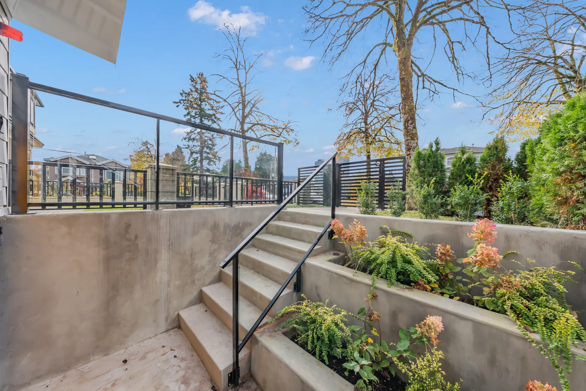 Concrete steps up from the patio to the front yard between planted walls, a glass railing at the top